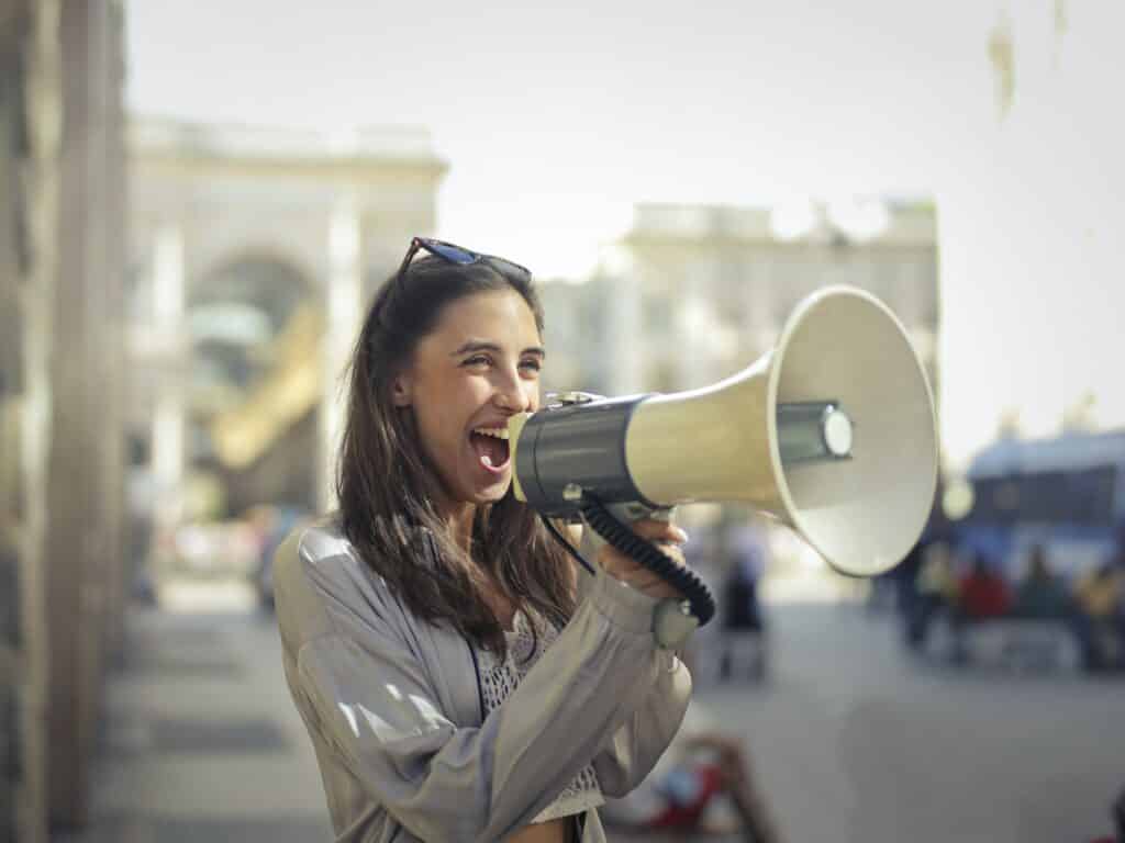 A young woman standing on the street side is shouting on a megaphone.