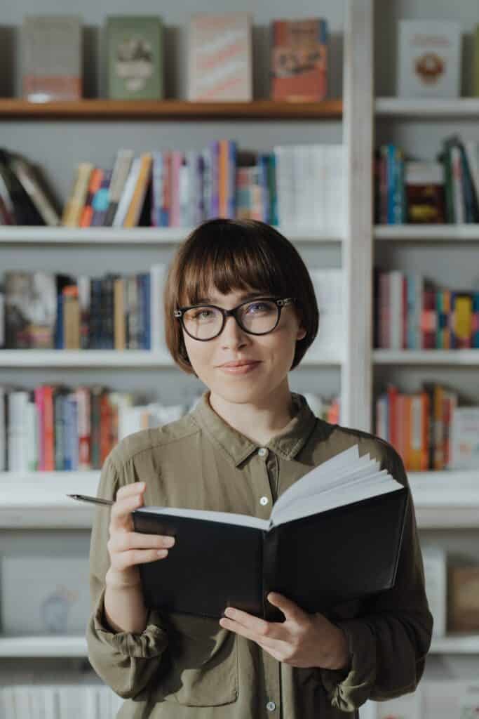 A happy lady holds a notebook and a pen while standing in front of a bookshelf.